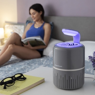 Dispositivo cinza escuro com luz azul sobre mesa branca, ao lado de óculos e livro, mulher ao fundo sentada no sofá