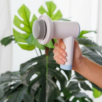 Aparelho massageador portátil branco com detalhes azuis segurado por mão em frente a folhas verdes