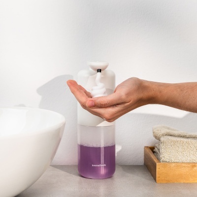 Frasco doseador com líquido roxo e tampa branca ao lado de uma pia branca, com mão a dispensar espuma e caixa de madeira com toalhas