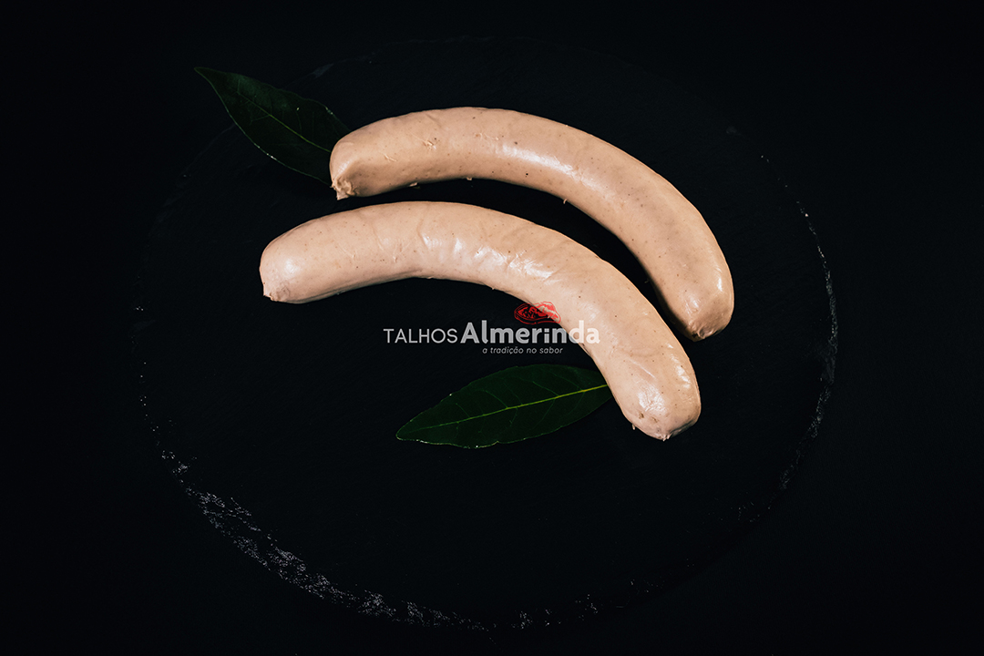 Salsicha Bratwurst Duas salsichas crus em prato preto com folhas verdes e texto TALHOS Almerinda