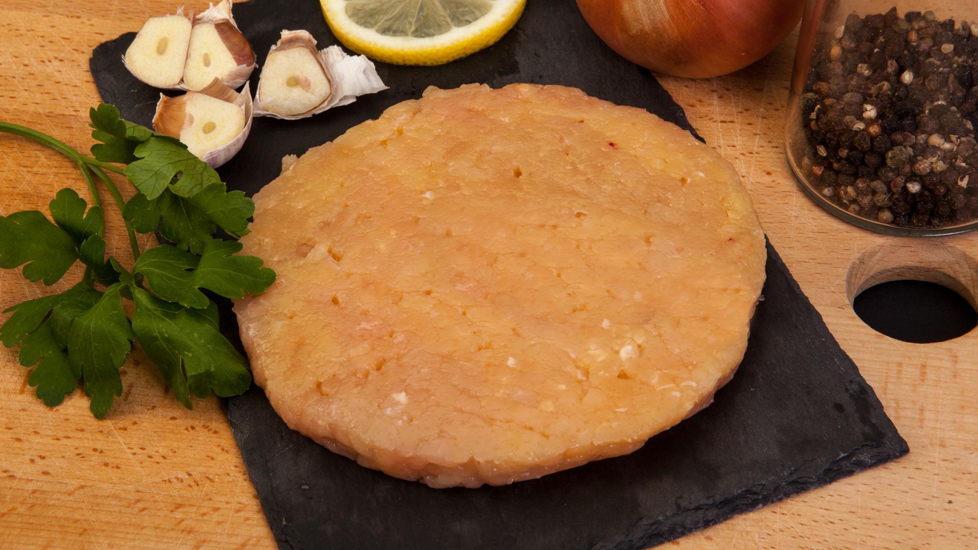 Hambúrguer de Frango do Campo 200g. Hambúrguer de frango cru redondo em tábua preta com alho, limão, salsa, cebola e pimenta-do-reino