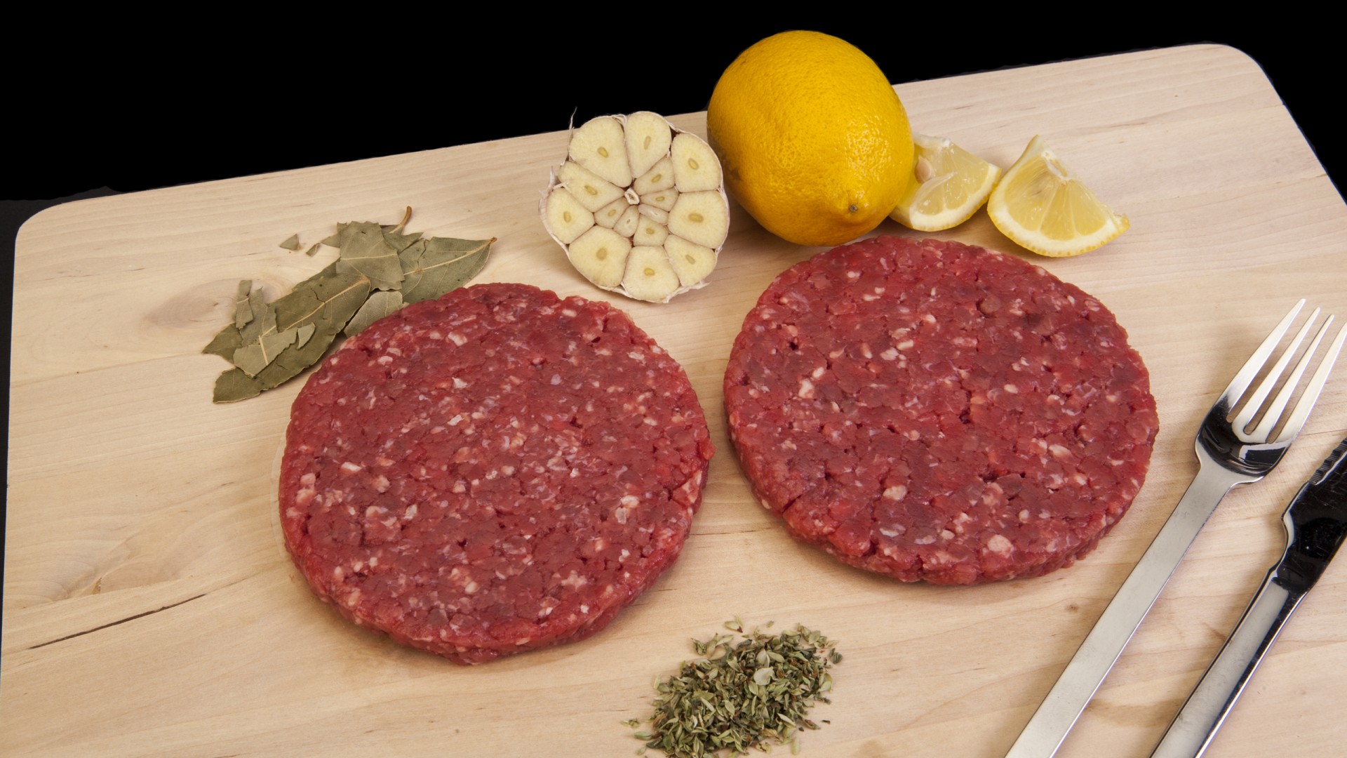Hambúrguer de Vaca 200g Hambúrgueres crus numa tábua com limão, alho, ervas, faca e garfo