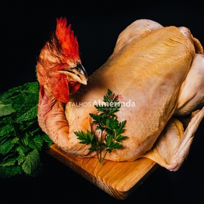 Frango inteiro cru com pele amarelada sobre tábua de madeira com ervas e fundo preto