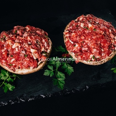 Dois cogumelos recheados com carne moída vermelha e pedaços brancos sobre base preta com salsa verde