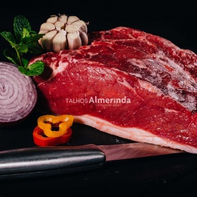 Pedaço de carne crua vermelho com alho, cebola roxa, pimentos e folhas verdes em superfície preta com faca