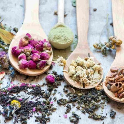 Colheres de pau com várias ervas e flores secas coloridas em superfície clara.