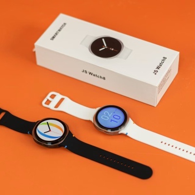 Dois relógios inteligentes JS Watch8 com braceletes preto e branco ao lado da sua caixa branca sobre fundo laranja