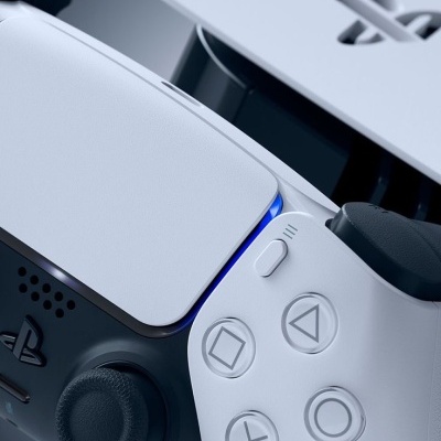 Comando de videojogo PlayStation 5 branco com símbolos nos botões e consola ao fundo