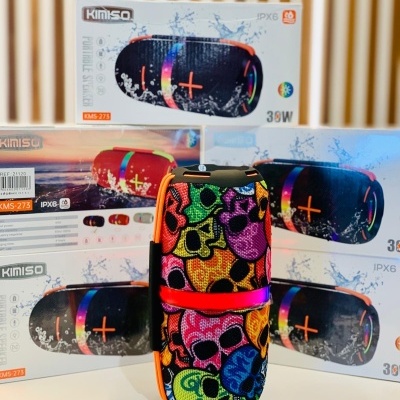 Coluna portátil Kimiso colorida com padrão de caveiras e caixas embalagens ao fundo.