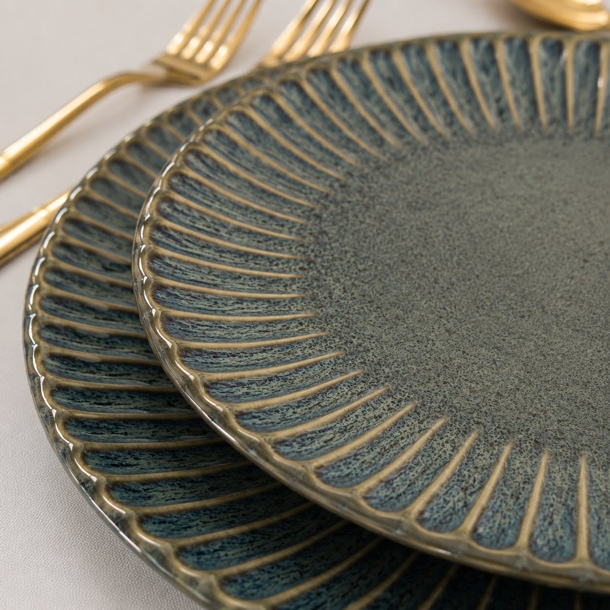 Conjunto de Louça de Mesa Azul Petróleo com Relevo Dois pratos de cerâmica azul-esverdeada com detalhes dourados e talheres dourados numa mesa clara