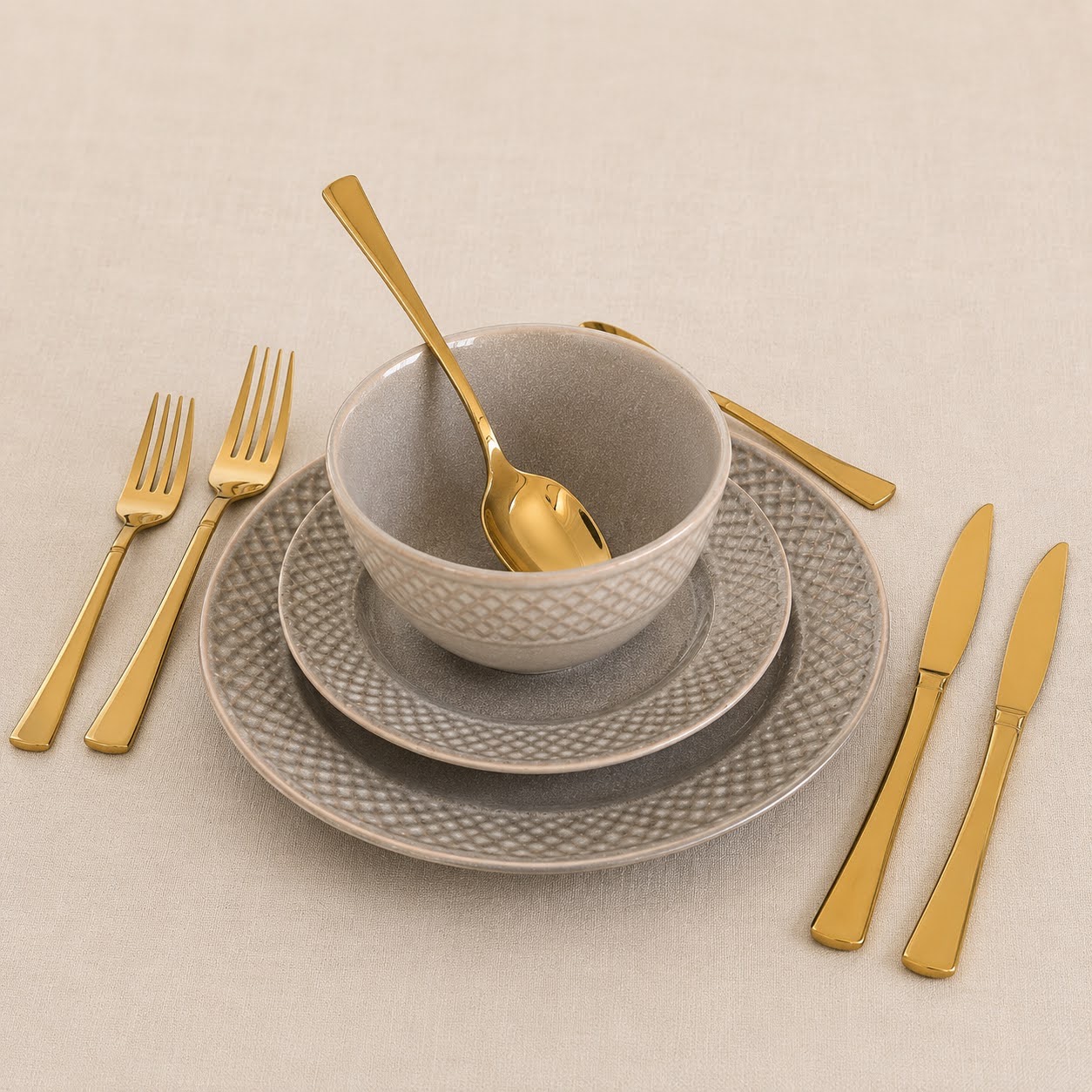 Conjunto de Louça de Mesa Bege Pérola em Grés Conjunto de loiça cinzenta com talheres dourados sobre superfície bege.