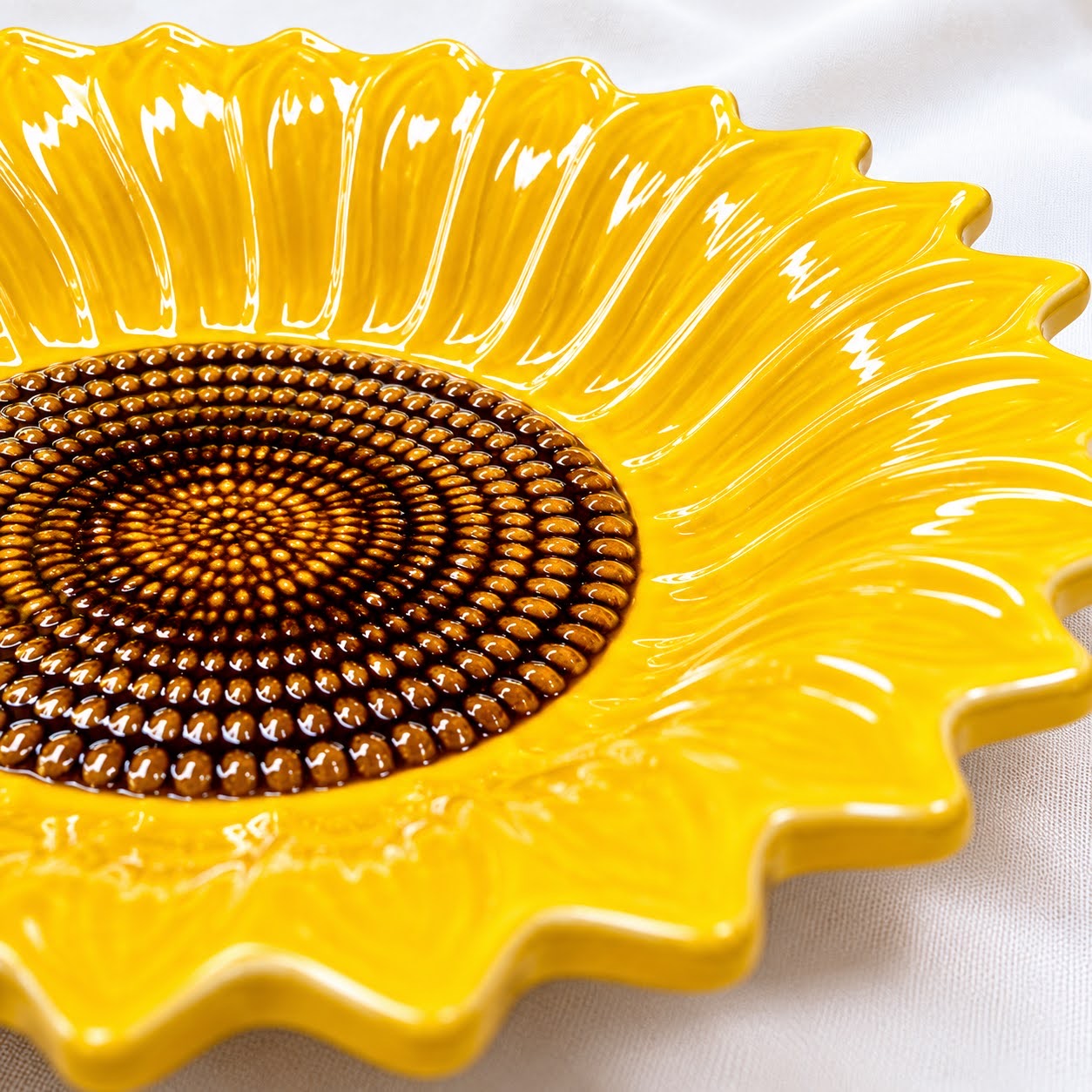 Coleção Girassóis Prato cerâmico amarelo em formato de girassol com centro marrom, sobre tecido branco.