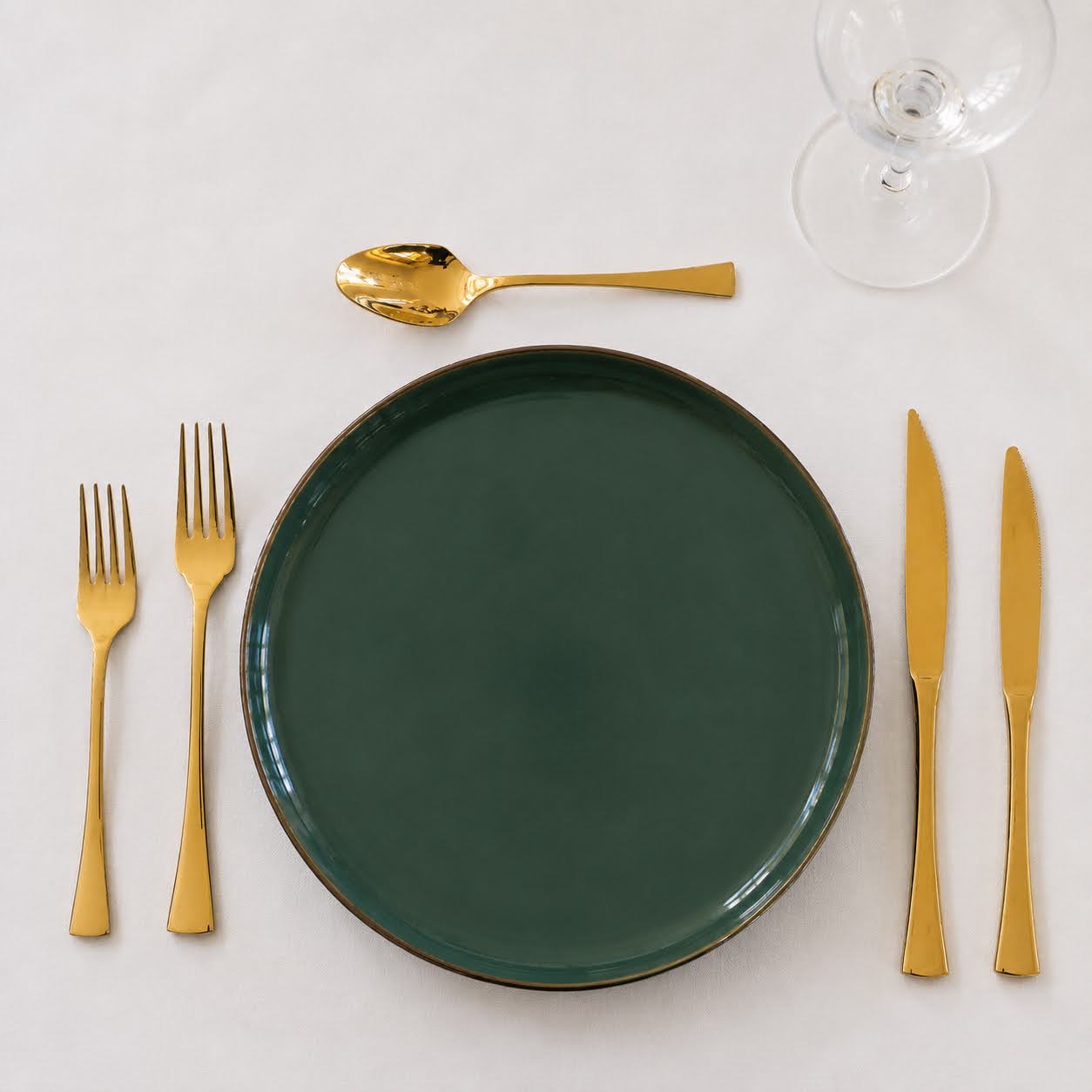 Conjunto de Mesa Verde Esmeralda | 3 Peças Talheres dourados ao redor de prato verde escuro com copo de vidro em fundo branco