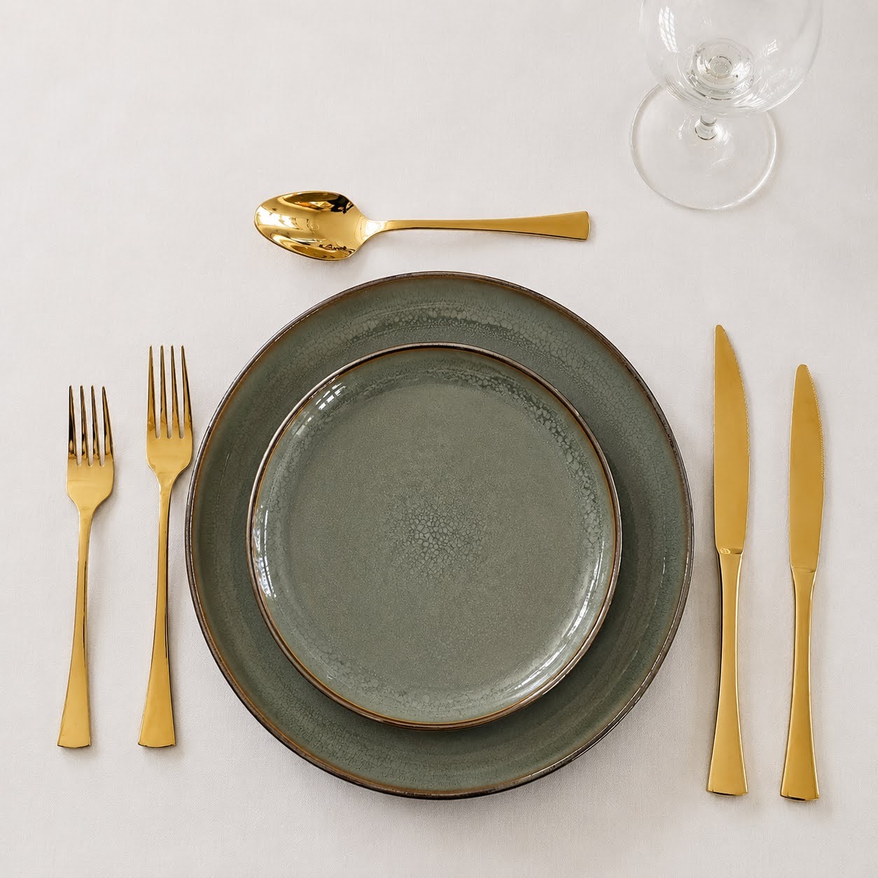 Conjunto de Mesa Verde | 3 Peças Conjunto de pratos cinzentos com talheres dourados e copo em toalha branca.