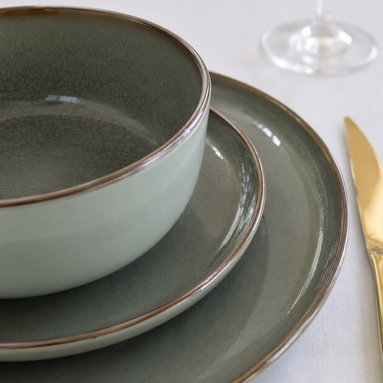 Conjunto de Mesa Verde | 3 Peças Conjunto de loiça verde com prato fundo, prato raso e prato de sobremesa sobre toalha branca com faca dourada e base de copo transparente
