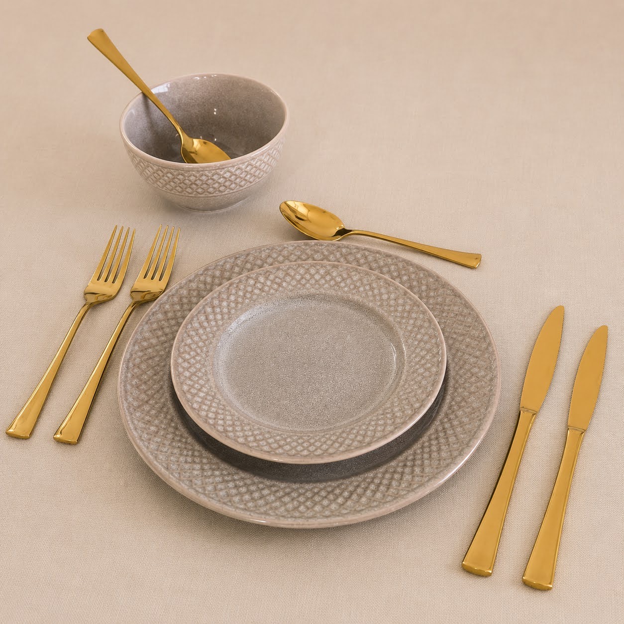 Conjunto de Louça de Mesa Bege Pérola em Grés Conjunto de loiça cinzenta com talheres dourados sobre tecido bege.