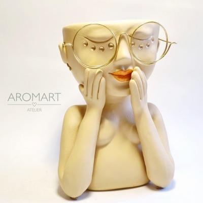 Escultura creme com rosto feminino e óculos dourados em fundo branco.