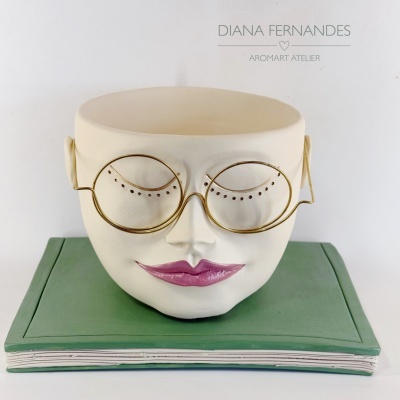 Vaso em forma de rosto branco com lábios rosa e óculos dourados sobre livro verde simulado