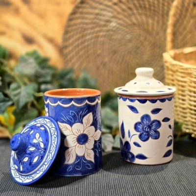 Dois frascos de cerâmica pintados à mão com flores, um azul e outro creme, com tampas, sobre mesa cinza e fundo folhas e vime.