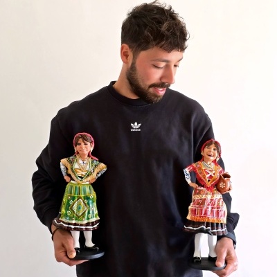 Homem com camisola preta segurando duas figuras em cerâmica vestidas com trajes tradicionais coloridos