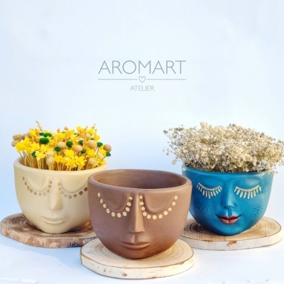 Três vasos cerâmicos com faces humanas e flores, sobre bases de madeira, com texto AROMART ATELIER no fundo.