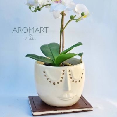 Vaso de cerâmica com rosto estilizado, orquídea branca, livro castanho e texto Aromart Atelier