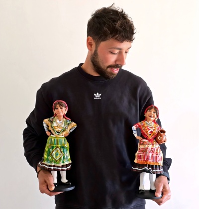 Homem com camisola preta segurando duas figuras em cerâmica vestidas com trajes tradicionais coloridos