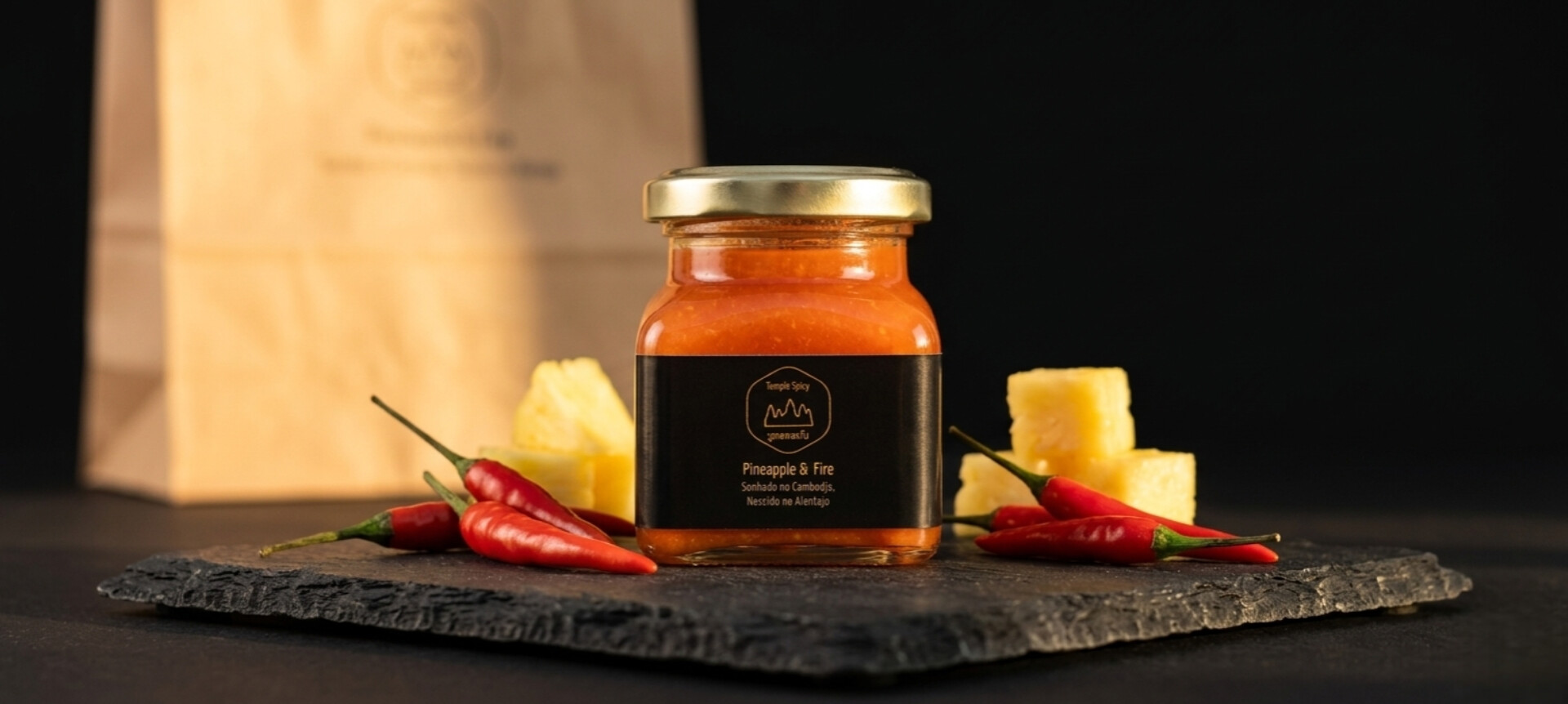 Pineapple & Fire - Picante com Abacaxi 100ml Frasco de pasta laranja com etiqueta preta, malaguetas e pedaços de ananás sobre pedra preta