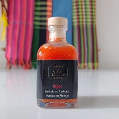 Garrafa de vidro com molho vermelho e etiqueta preta com texto, sobre superfície branca e tecidos coloridos ao fundo