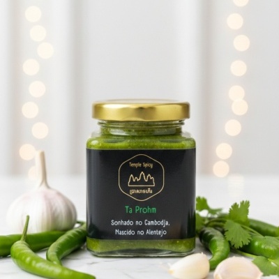 Frasco de pasta verde com etiqueta preta e tampa dourada rodeado por malaguetas, alho e coentros.