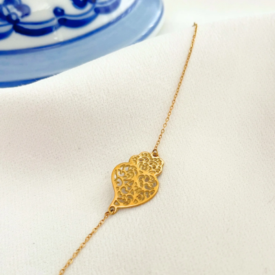 Pulseira dourada com pendente em forma de coração com padrão recortado sobre pano branco e prato azul e branco ao fundo