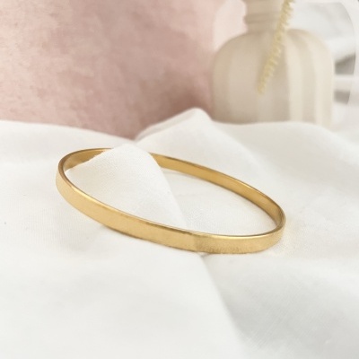 Pulseira dourada lisa sobre tecido branco com fundo rosa e objeto decorativo ao fundo