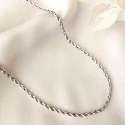 Colar prateado de corrente torcida sobre fundo branco com decoração floral clara