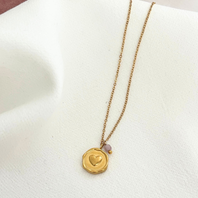 Colar dourado com pendente redondo com coração e pedra rosa sobre tecido branco