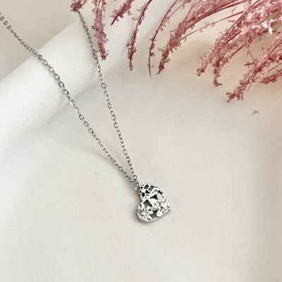 Colar prateado com pendente de coração florido sobre fundo branco e flores secas cor de rosa