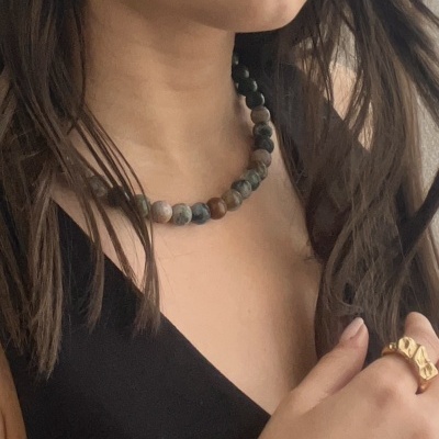 Mulher com colar de contas redondas coloridas e foscas, brinco dourado texturizado e anel dourado geométrico.