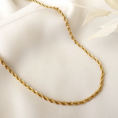 Colar dourado de corrente torcida sobre tecido branco com folha seca