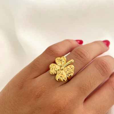 Anel dourado com quatro folhas em forma de coração numa mão com unhas vermelhas.