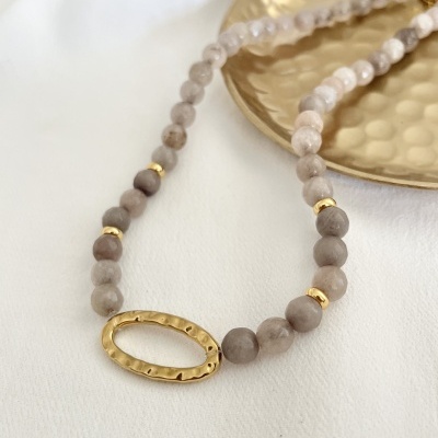 Colar de contas cinza e bege com pendente dourado oval sobre tecido branco e prato dourado