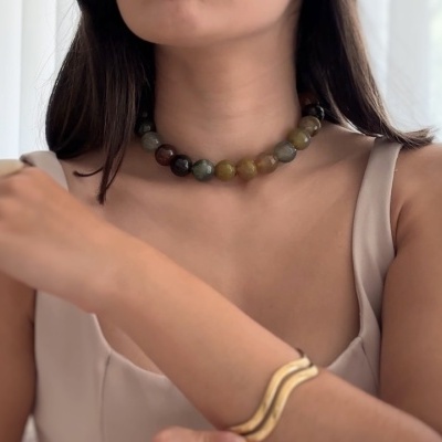 Colar de contas coloridas com pulseira dourada, usado por mulher com vestido bege.