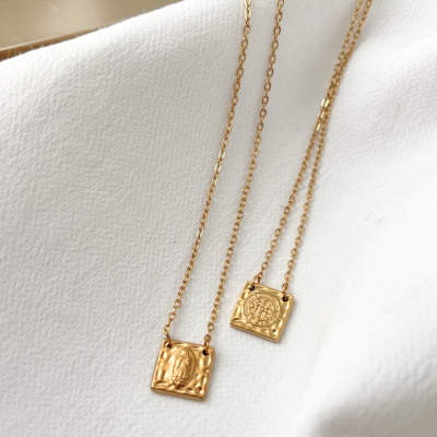 Colares dourados com pendentes quadrados texturizados sobre tecido branco.