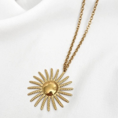 Colar dourado com pendente em forma de sol sobre tecido branco.