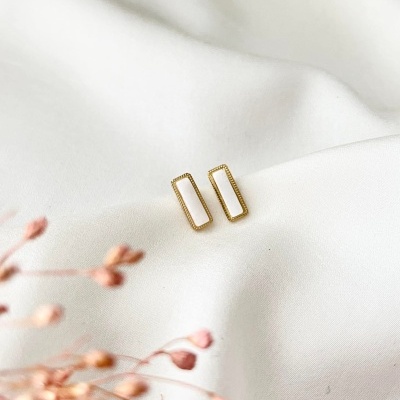 Brincos retangulares dourados com centro branco sobre tecido branco e flores rosa desfocadas