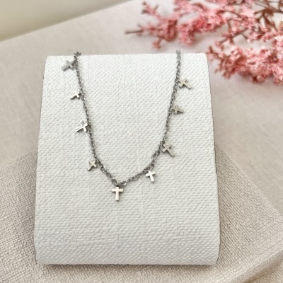 Colar prateado com vários pequenos pingentes em forma de cruz num suporte branco texturizado