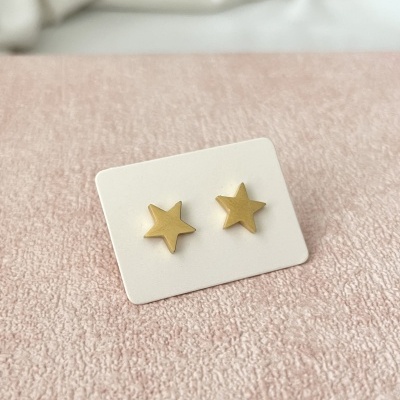 Brincos estrela dourados em base branca sobre tecido rosa.