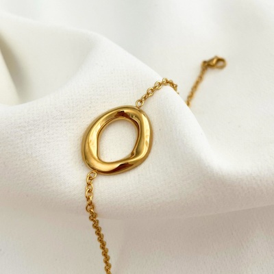 Pulseira dourada com pendente oval irregular sobre tecido branco