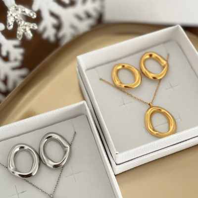 Conjuntos de joias ovais em prata e dourado em caixas brancas sobre base dourada com decoração natalícia e laço cor-de-rosa ao fundo.