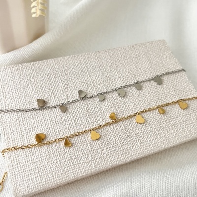Duas pulseiras de correntes com pendentes de corações, uma prateada e outra dourada, sobre suporte claro texturado.