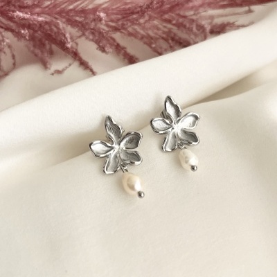 Brincos de flor de metal prateado com pérola pendurada sobre tecido branco com folhagem rosa ao fundo