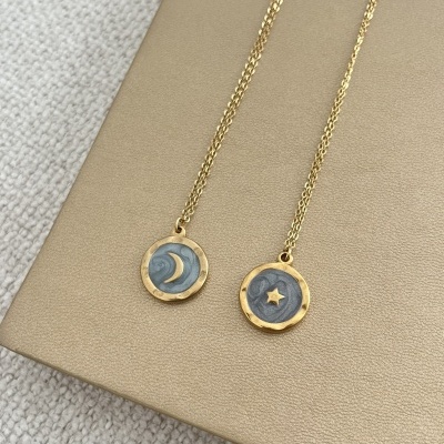 Dois colares dourados com pendentes de lua e estrela sobre fundo bege.